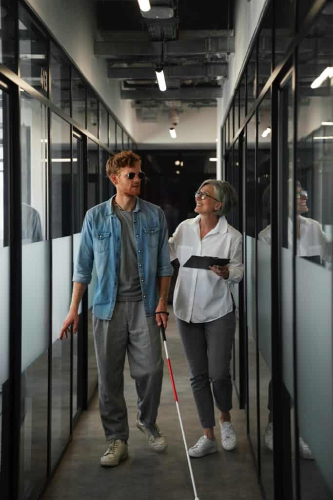 Dans un couloir, un jeune homme marche en utilisant sa canne est accompagné de sa collègue plus âgée 